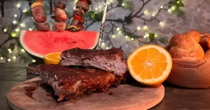 Barbecue ribs presented next to an orange, a bowl of buns, and meat kebabs stuck in a watermelon slice