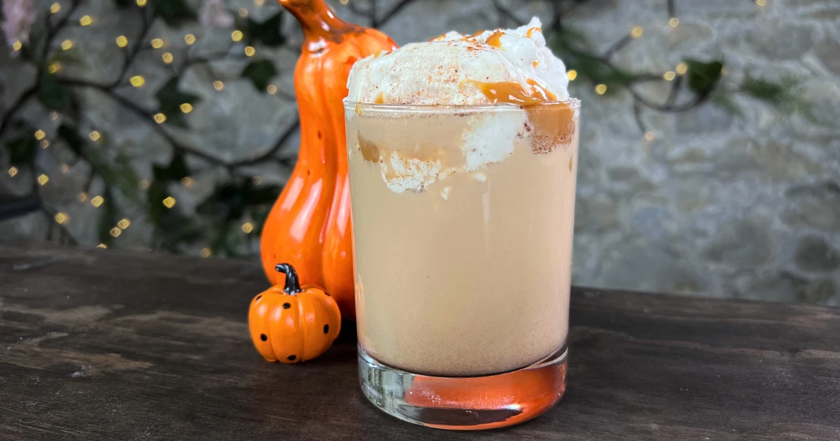 A pumpkin spice, dulce de leche flavored drink in a clear glass with two decorative pumpkins behind it