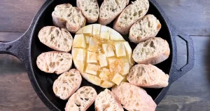Baked Honey and Butter Cheese Wheel in a cast iron pan, surrounded by baguette slices
