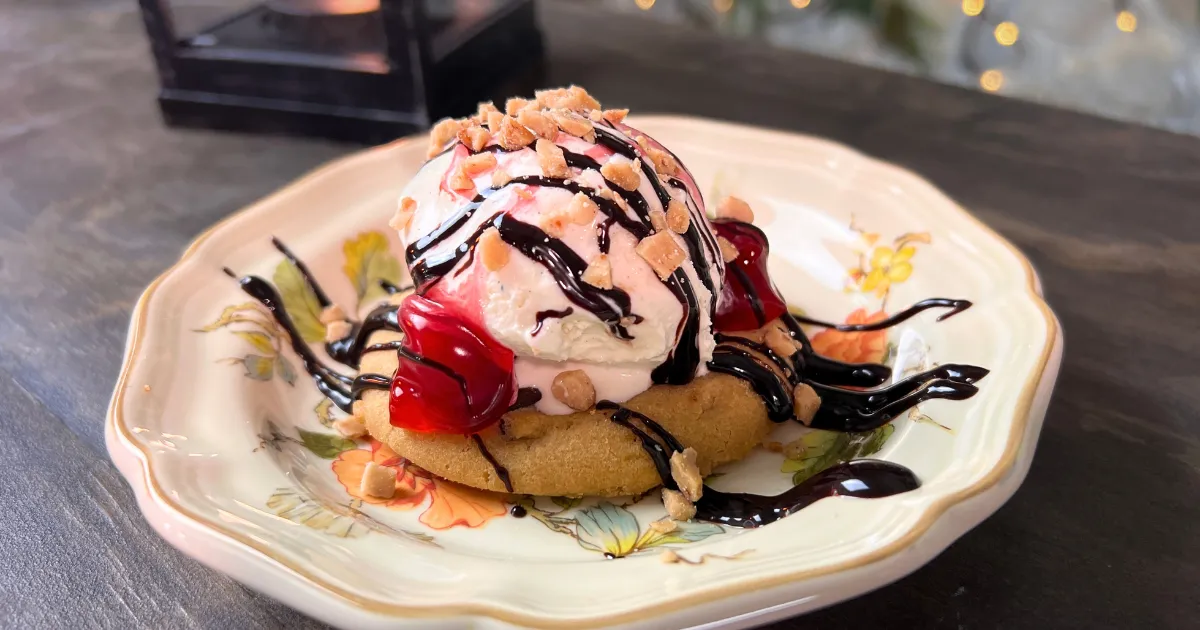 A cookie on a plate topped with ice cream, cherries, toffee, and chocolate syrup.